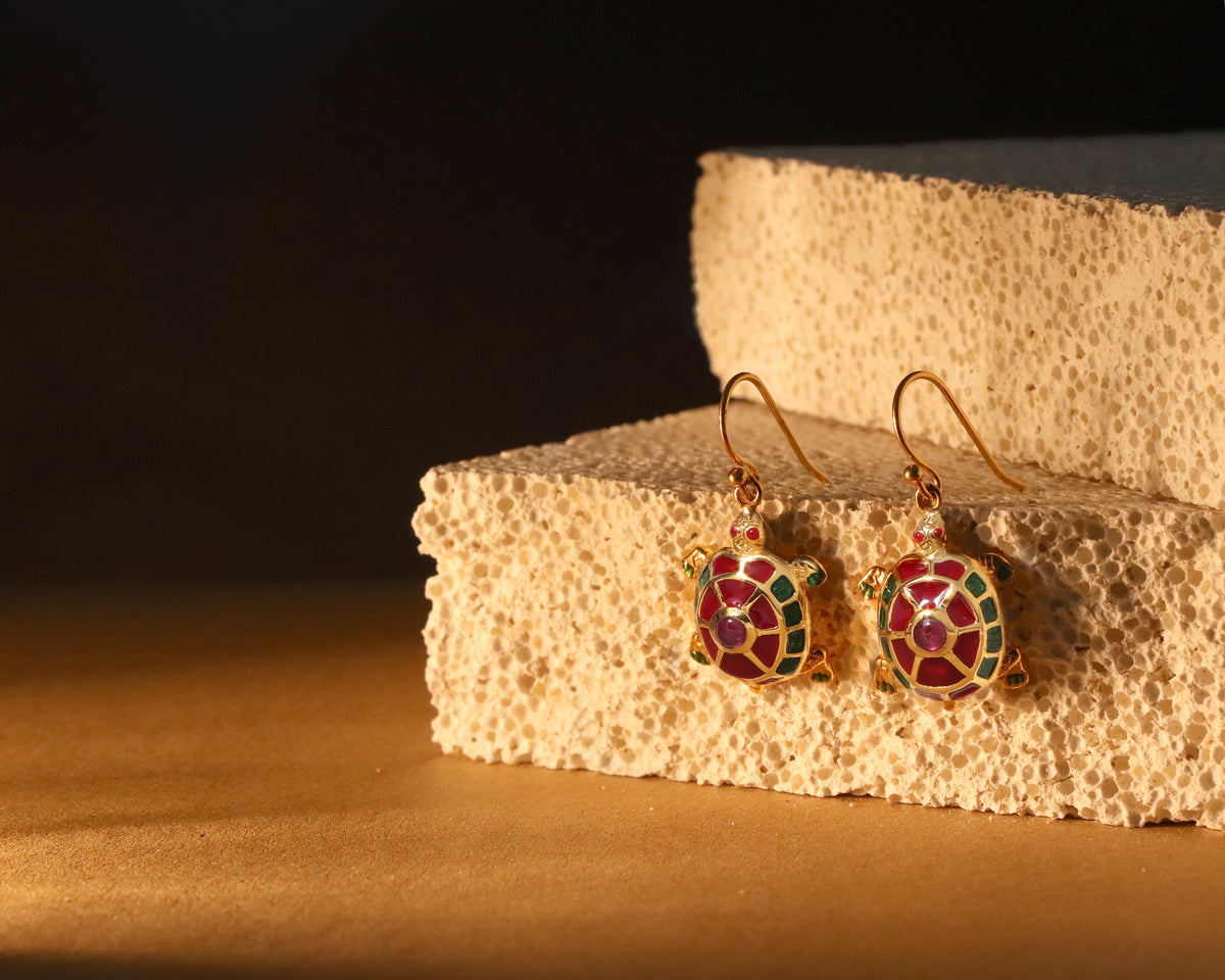A pair of gold-tone turtle drop earrings with red, green, and blue enamel detailing, displayed on a textured beige stone block against a dark background.