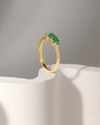 A gold ring with a cluster of green gemstones, displayed on a white sculptural pedestal against a neutral gray background.