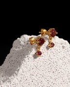 A pair of gold drop earrings with oval red gemstones, displayed on a textured white stone pedestal against a solid black background.