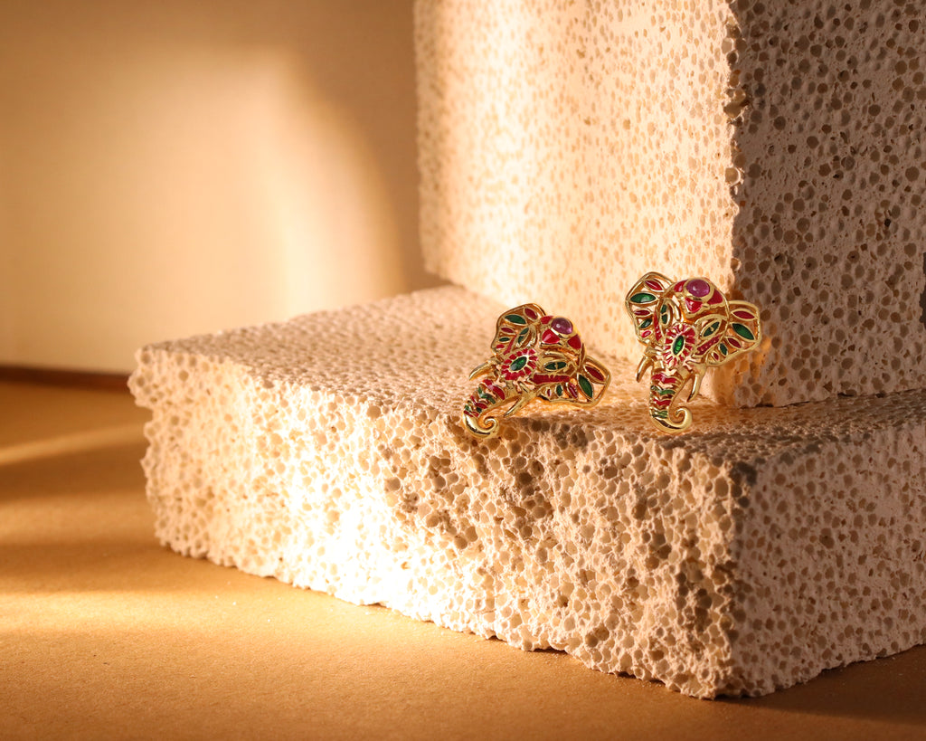 A pair of gold-toned elephant stud earrings decorated with red and green enamel, displayed on a textured beige foam block under warm, directional lighting.
