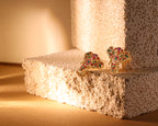 A pair of gold-toned elephant stud earrings decorated with red and green enamel, displayed on a textured beige foam block under warm, directional lighting.