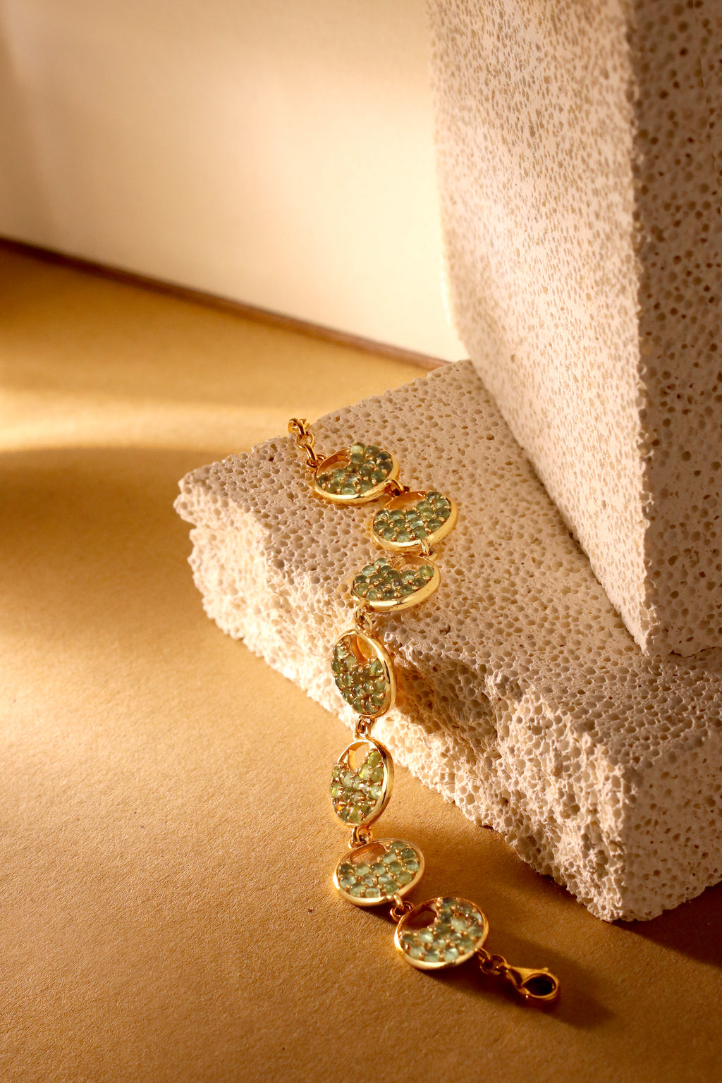 A gold link bracelet with oval green stone inlays draped over a textured beige stone block on a warm beige surface.
