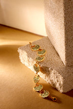 A gold link bracelet with oval green stone inlays draped over a textured beige stone block on a warm beige surface.