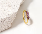 A gold ring with a cluster of purple gemstones, photographed on a white surface next to a textured white stone prop.