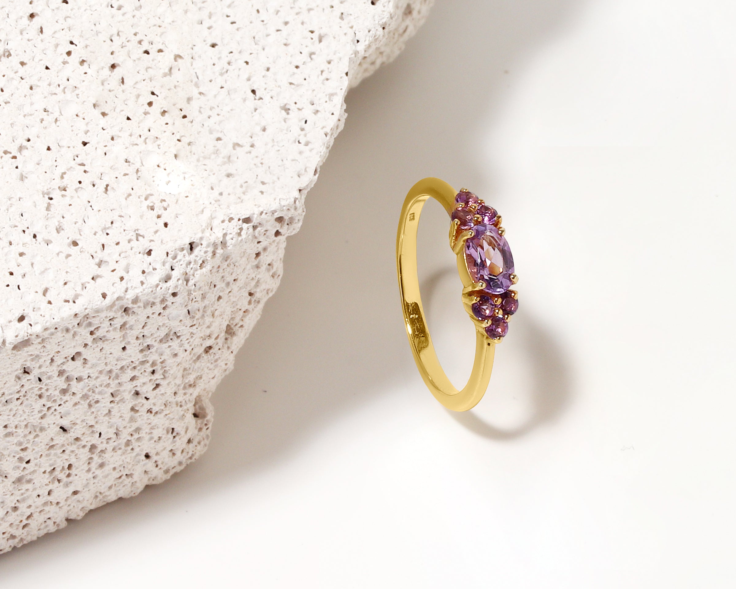 A gold ring with a cluster of purple gemstones, photographed on a white surface next to a textured white stone prop.
