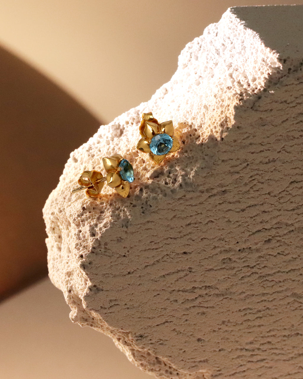 A pair of gold floral stud earrings with light blue gemstones, displayed on a textured beige stone block against a warm beige background.