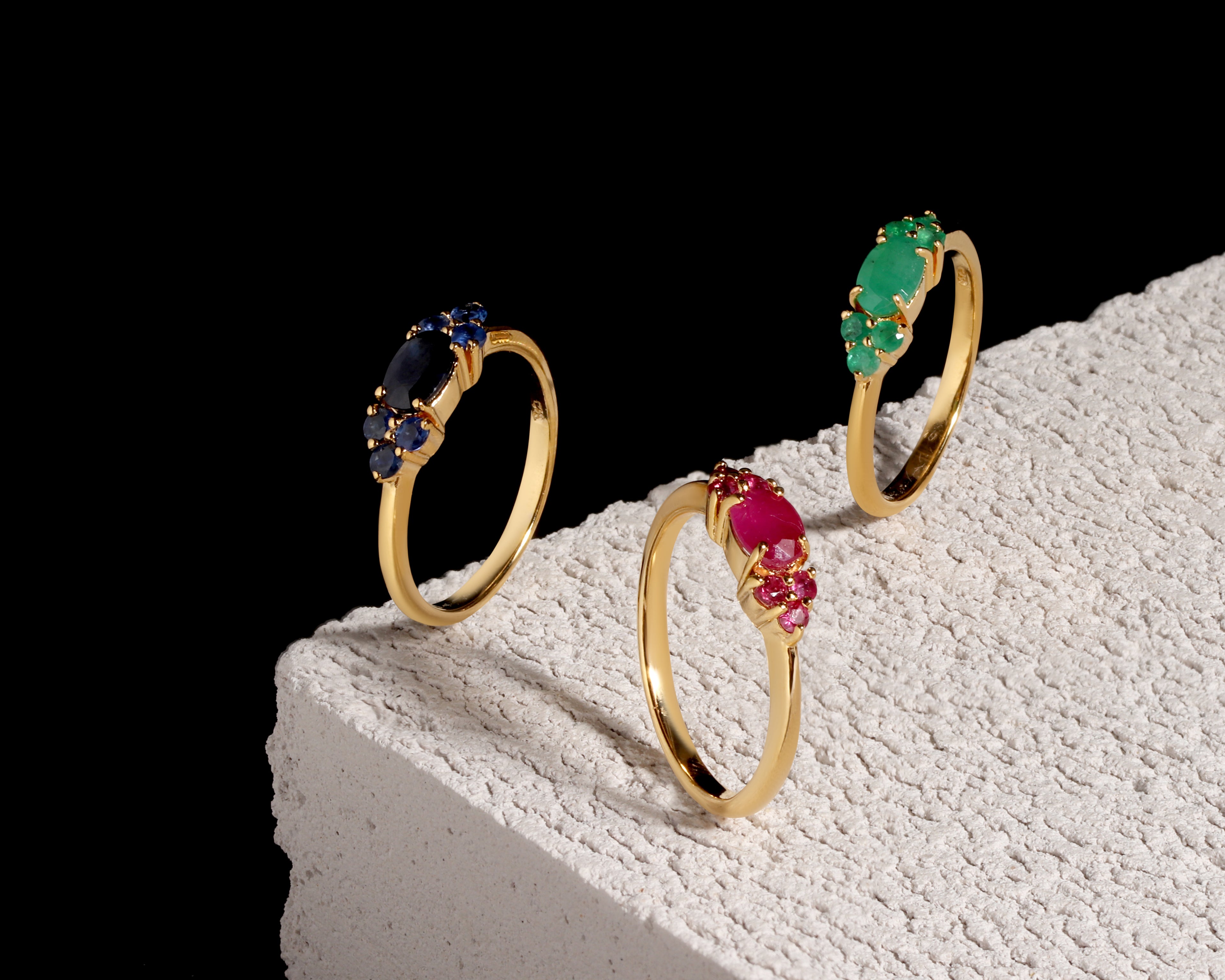 Three gold rings with marquise-cut gemstones in blue, green, and pink, displayed on a textured white block against a black background.