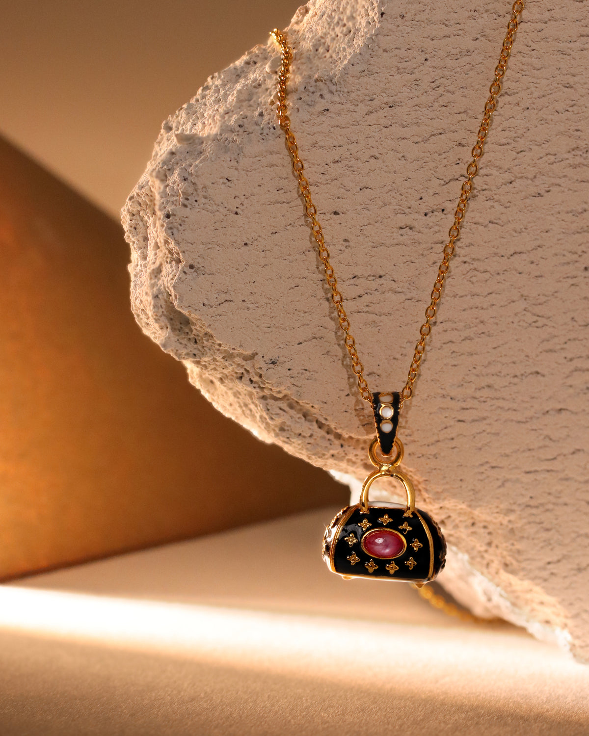 A gold-tone necklace with a handbag-shaped pendant, featuring black enamel, gold floral details, and a central pink gemstone, displayed on a textured stone prop with warm, dramatic lighting.