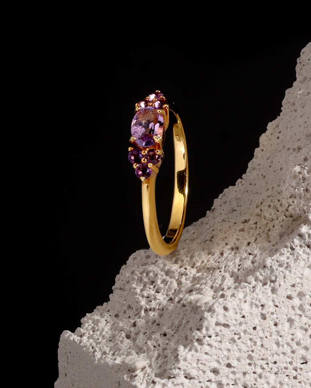 A yellow gold ring with a marquise-cut purple amethyst center stone and smaller round purple accent stones, displayed on a textured white stone prop against a solid black background.