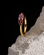 A yellow gold ring with a marquise-cut purple amethyst center stone and smaller round purple accent stones, displayed on a textured white stone prop against a solid black background.