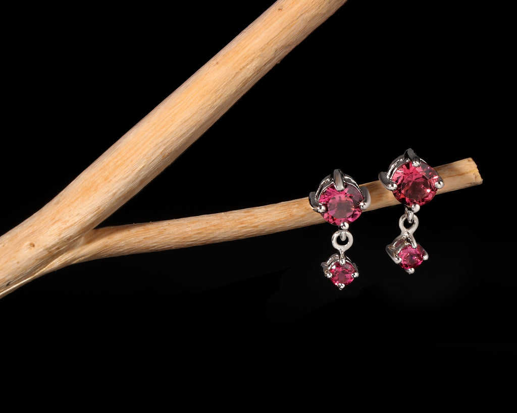 A pair of silver-toned dangle earrings with round and rectangular pink gemstones, displayed on a light wood branch against a solid black background.