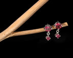 A pair of silver-toned dangle earrings with round and rectangular pink gemstones, displayed on a light wood branch against a solid black background.