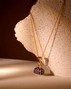 A gold chain necklace with a handbag-shaped pendant, decorated with blue, red, and gold enamel, displayed on a textured stone prop against a dark brown background.