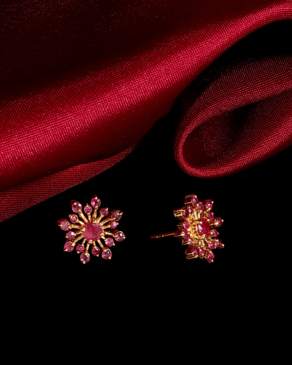A pair of gold-toned floral stud earrings with pink gemstones, displayed on a black background with draped red satin fabric.