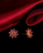 A pair of gold-toned floral stud earrings with pink gemstones, displayed on a black background with draped red satin fabric.