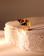 A gold ring featuring a sculpted black animal figure with a prominent red gemstone, displayed on a textured beige stone pedestal against a warm brown background.