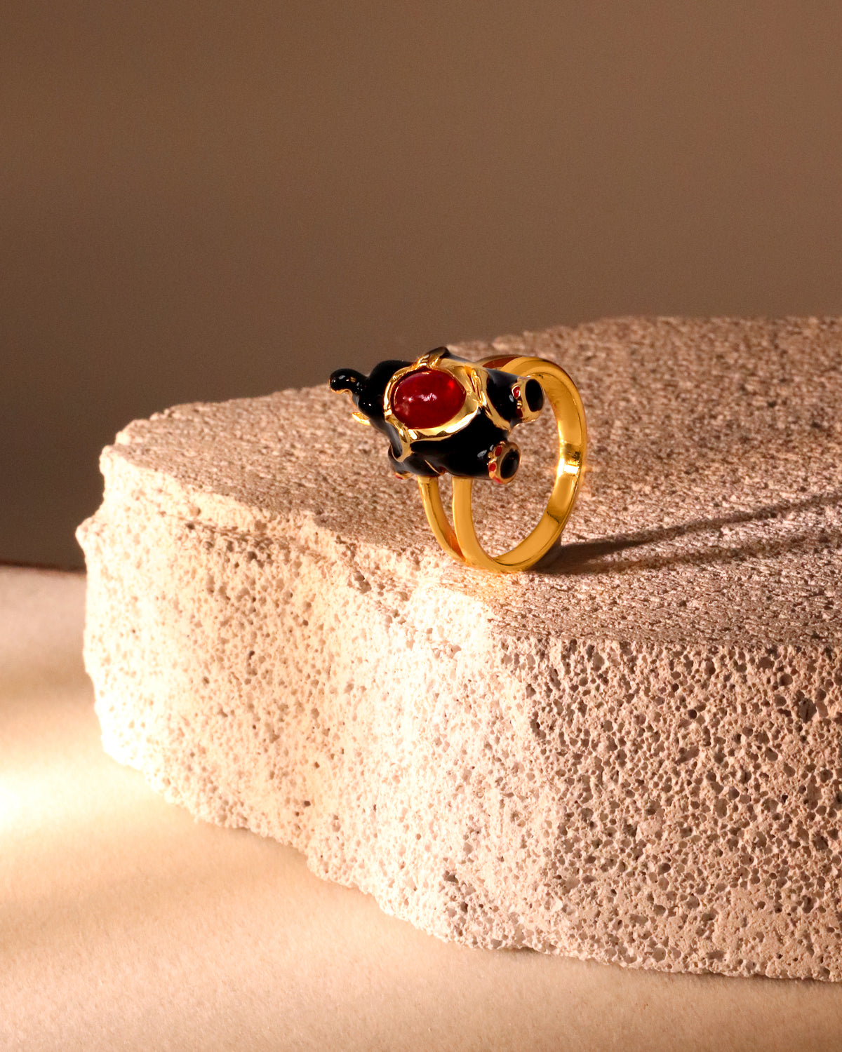 A gold ring featuring a sculpted black animal figure with a prominent red gemstone, displayed on a textured beige stone pedestal against a warm brown background.