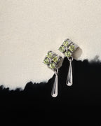A pair of silver drop earrings with four small light green gemstones in a square setting, each connected by a drop-shaped silver chain, displayed on a textured off-white surface with a black torn-paper accent.