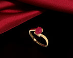 A gold ring with an oval pinkish-red gemstone, displayed on a black background with a draped red satin fabric.