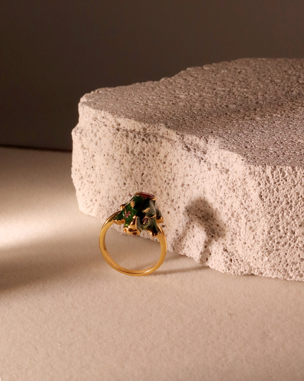 A gold ring with a green and red floral crown setting, resting against a textured beige stone block on a neutral beige surface.