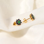 A pair of gold flower stud earrings with green center stones and dark blue outer stones, displayed on a soft white fabric background.