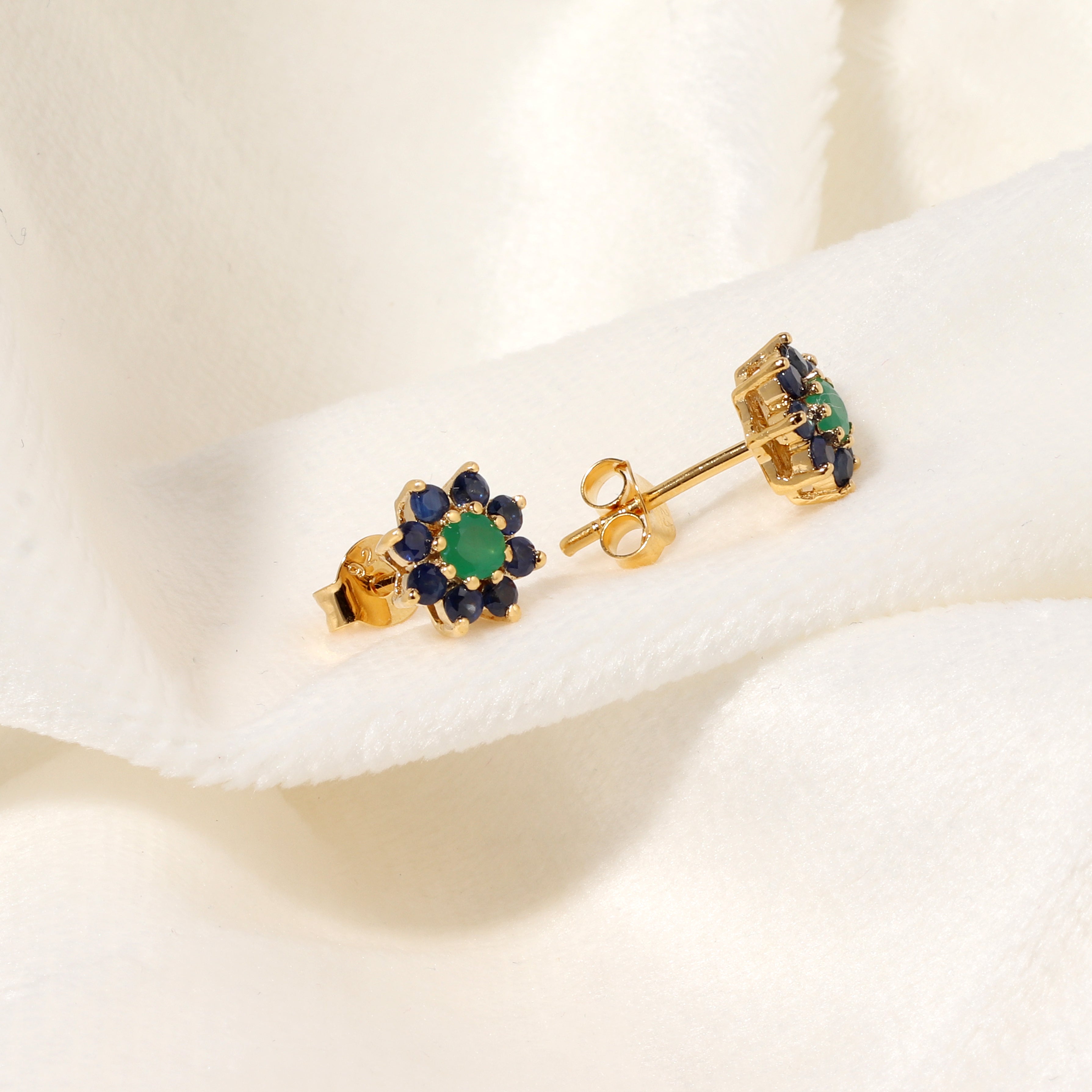 A pair of gold flower stud earrings with green center stones and dark blue outer stones, displayed on a soft white fabric background.