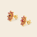 A pair of gold flower-shaped stud earrings with red gemstone centers and surrounding pinkish-red stones, displayed on a light beige background.