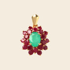 A gold pendant featuring a central oval-cut green emerald gemstone surrounded by a halo of round deep red ruby gemstones, set in a gold frame against a light beige background.
