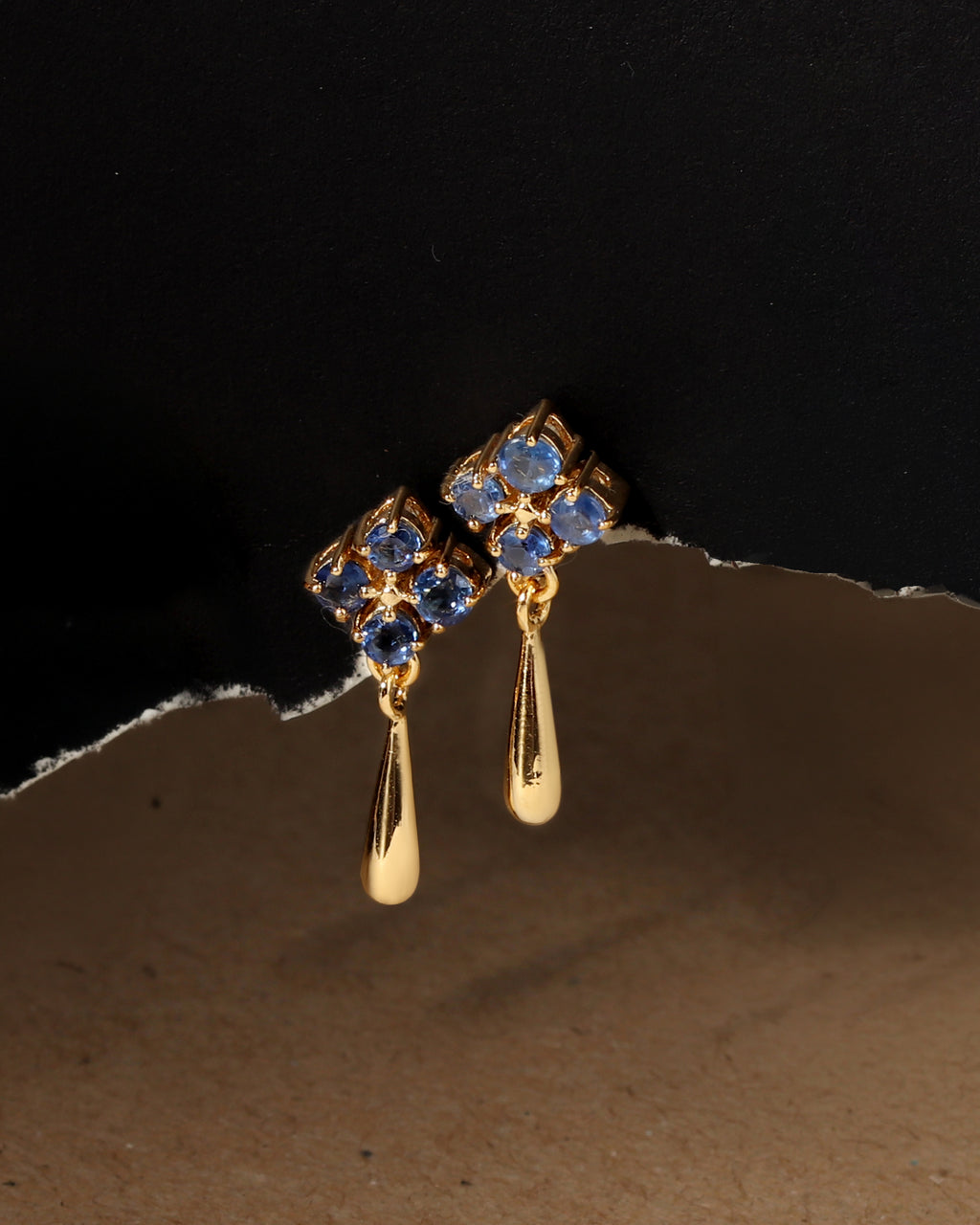 A pair of gold dangle earrings with blue gemstone floral clusters and teardrop-shaped drops, photographed against a dark background with a torn-paper style divider.