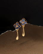 A pair of gold dangle earrings with blue gemstone floral clusters and teardrop-shaped drops, photographed against a dark background with a torn-paper style divider.