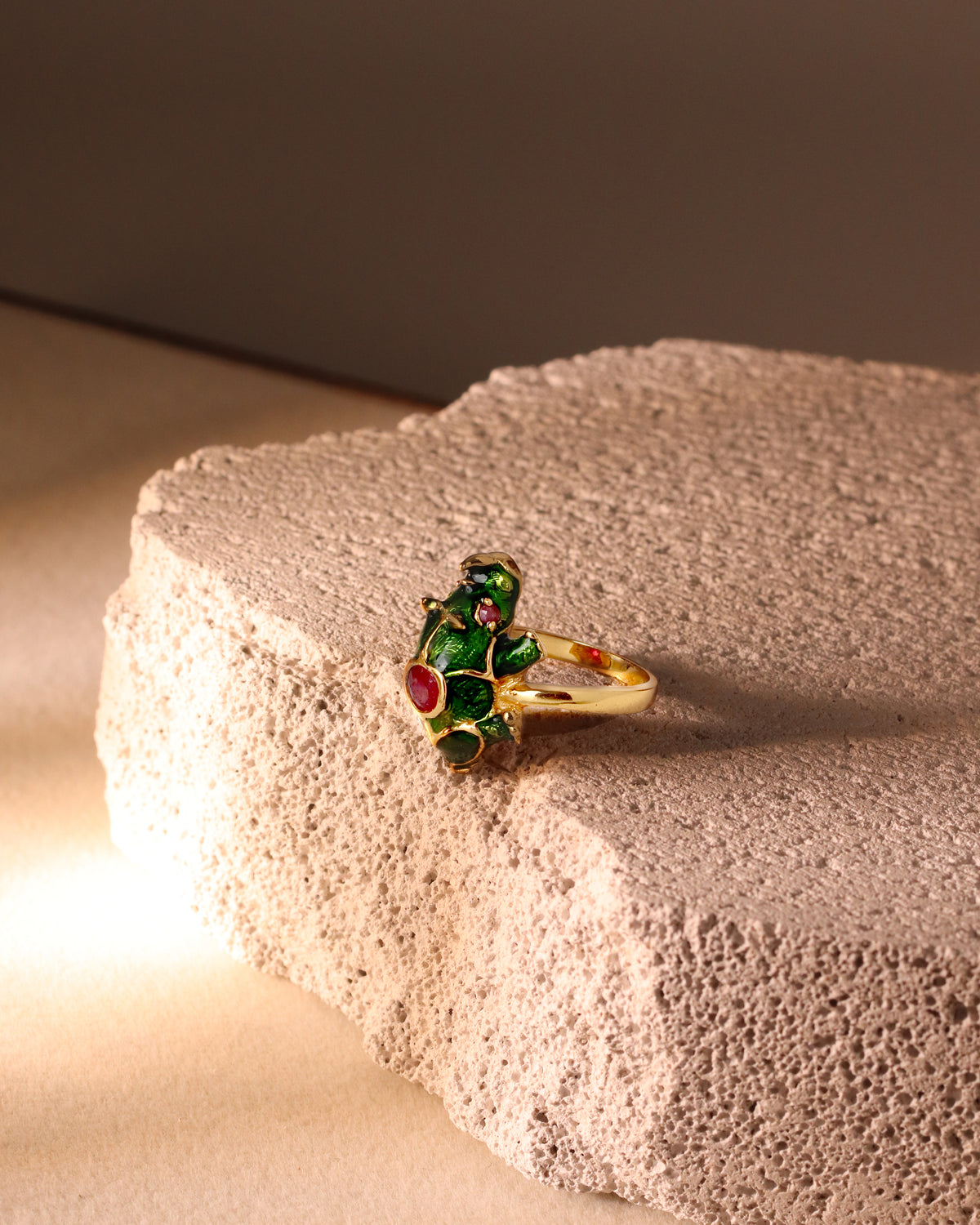 A gold ring featuring a frog-shaped setting with green enamel and red gemstone accents, displayed on a textured beige stone block.