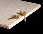 A pair of gold drop earrings with small green gemstone clusters, displayed on a textured beige stone slab against a solid black background.
