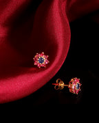 A pair of gold stud earrings with a central dark blue oval stone surrounded by small pink stones, displayed on a draped red satin fabric against a black background.