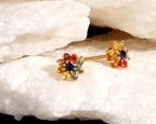 A pair of gold post earrings with multicolor gemstone clusters, displayed on a textured white rock surface.