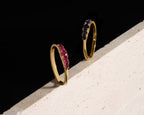 Two slim gold rings leaning against a textured white surface on a black background. One ring is set with bright pink gemstones, and the other is set with dark blue gemstones.