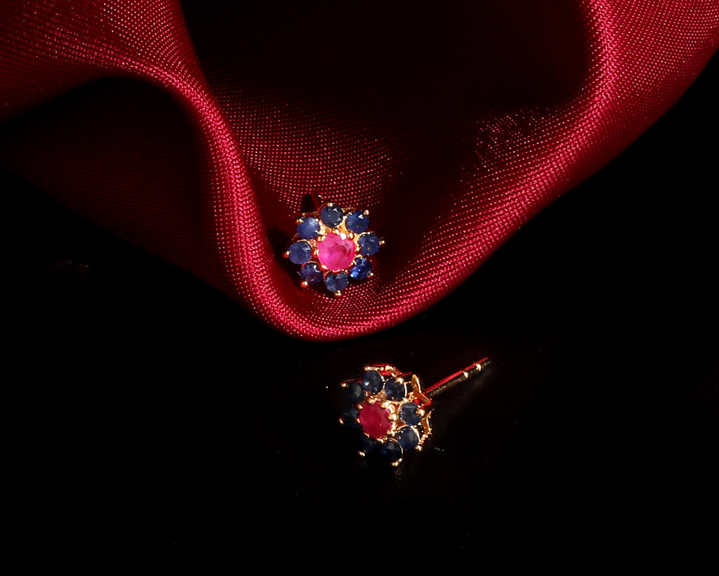A pair of gold-toned stud earrings with a central pink gemstone surrounded by small blue gemstones, displayed on a dark background with a draped red satin cloth.
