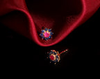 A pair of gold-toned stud earrings with a central pink gemstone surrounded by small blue gemstones, displayed on a dark background with a draped red satin cloth.