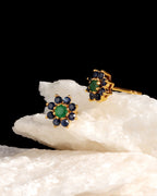 A pair of gold floral stud earrings with green center stones and dark blue surrounding stones, displayed on a white textured rock against a solid black background.