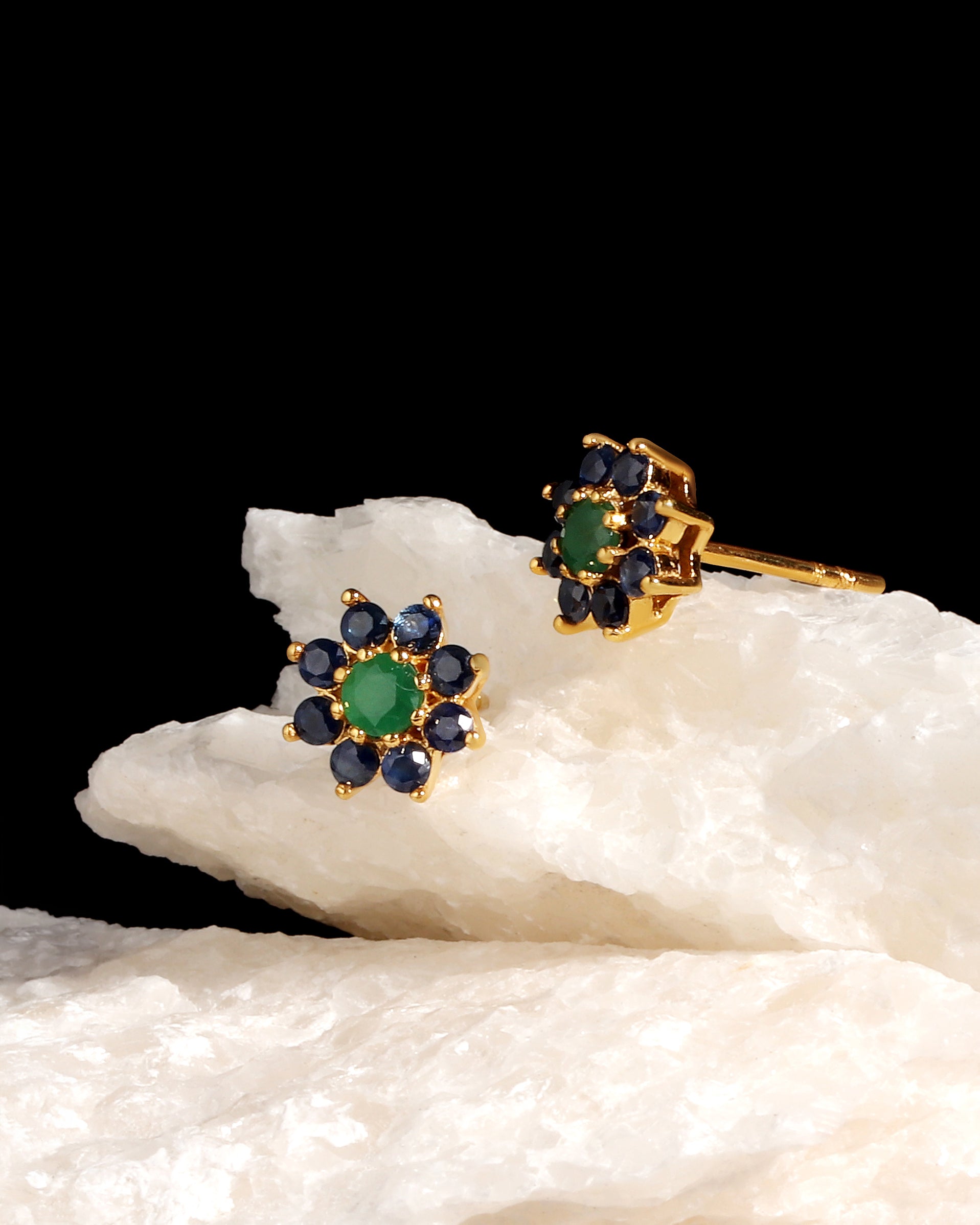 A pair of gold floral stud earrings with green center stones and dark blue surrounding stones, displayed on a white textured rock against a solid black background.