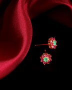 A pair of gold-toned stud earrings with bright green oval center stones surrounded by small pink stones, displayed on a black background with a draped red fabric accent.
