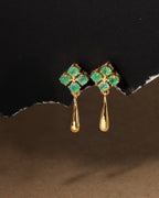 A pair of gold drop earrings with four green gemstones in a clover-like cluster at the top and a teardrop-shaped drop below, displayed against a dark textured background.