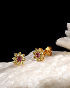 A pair of gold flower-shaped stud earrings with a central purple gemstone and surrounding clear stones, displayed on a white textured surface against a black background.