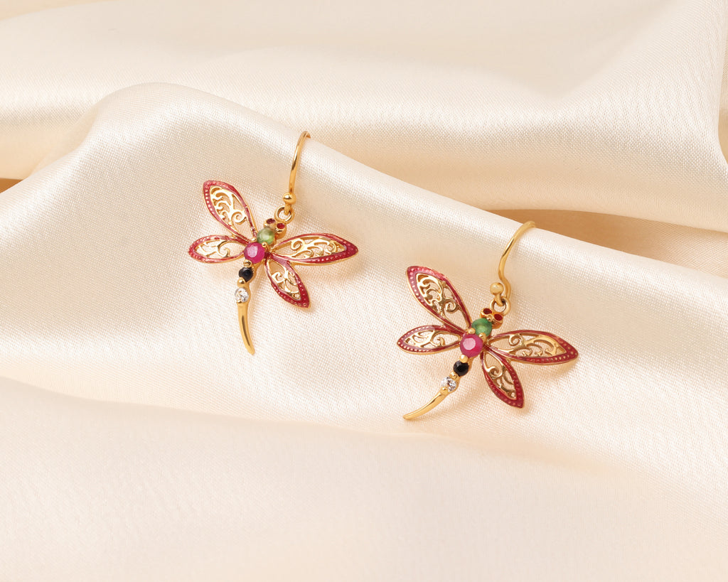 A pair of gold dragonfly earrings with red enamel wings and small colorful gemstone accents, displayed on draped cream satin fabric.
