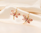 A pair of gold dragonfly earrings with red enamel wings and small colorful gemstone accents, displayed on draped cream satin fabric.