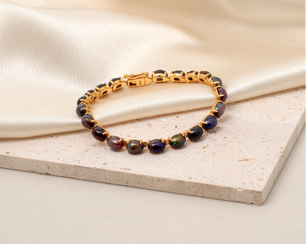 A gold-plated tennis bracelet with faceted, iridescent dark purple and green gemstones, resting on a textured beige stone slab with a draped cream satin fabric background.