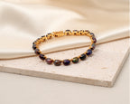 A gold-plated tennis bracelet with faceted, iridescent dark purple and green gemstones, resting on a textured beige stone slab with a draped cream satin fabric background.