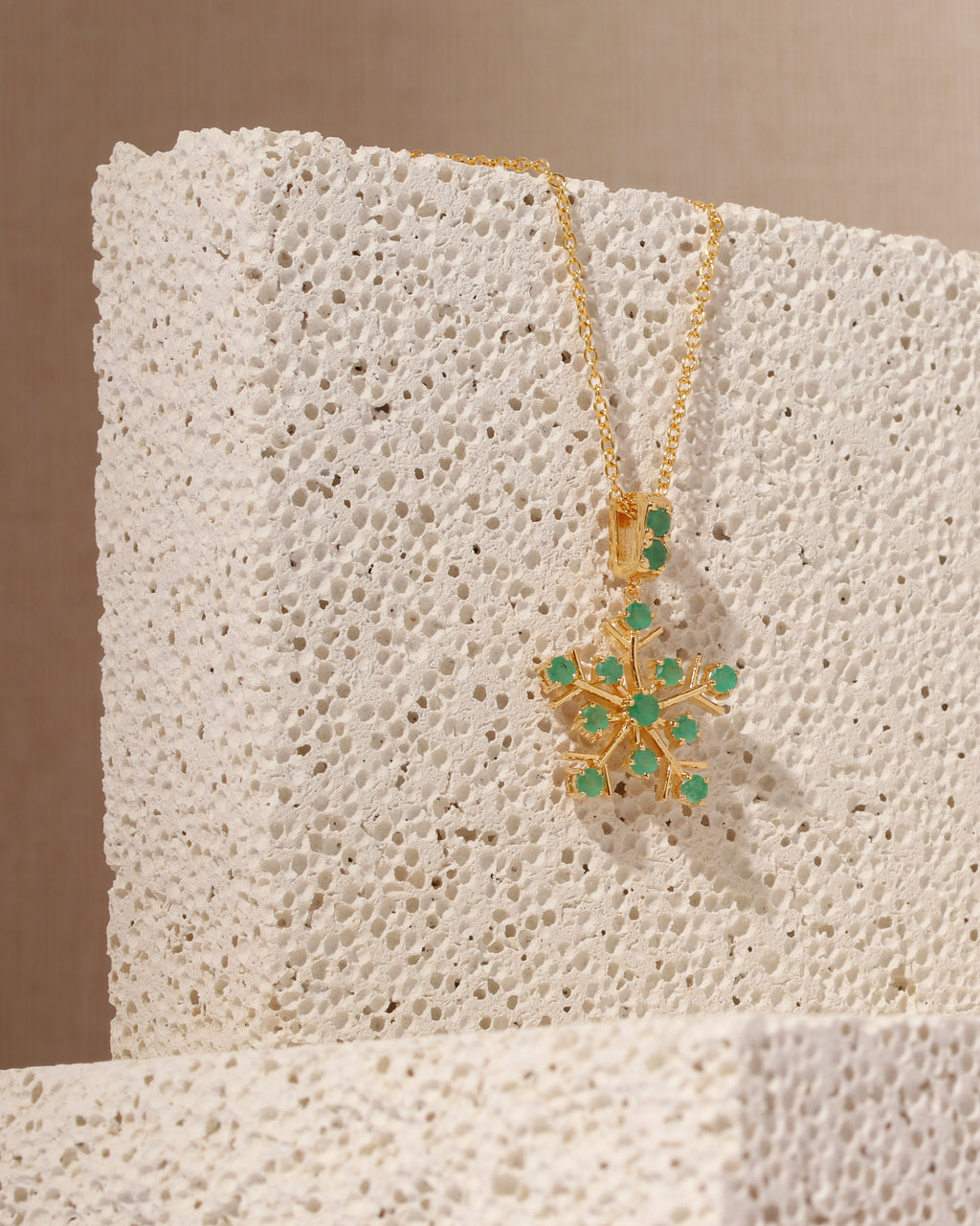 A gold pendant necklace with a snowflake design set with green gemstones, displayed on a textured white stone block against a warm beige background.