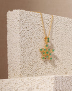 A gold pendant necklace with a snowflake design set with green gemstones, displayed on a textured white stone block against a warm beige background.