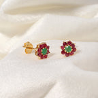 A pair of gold flower-shaped stud earrings with green oval center stones and surrounding rows of red round stones, displayed on a soft white fabric background.