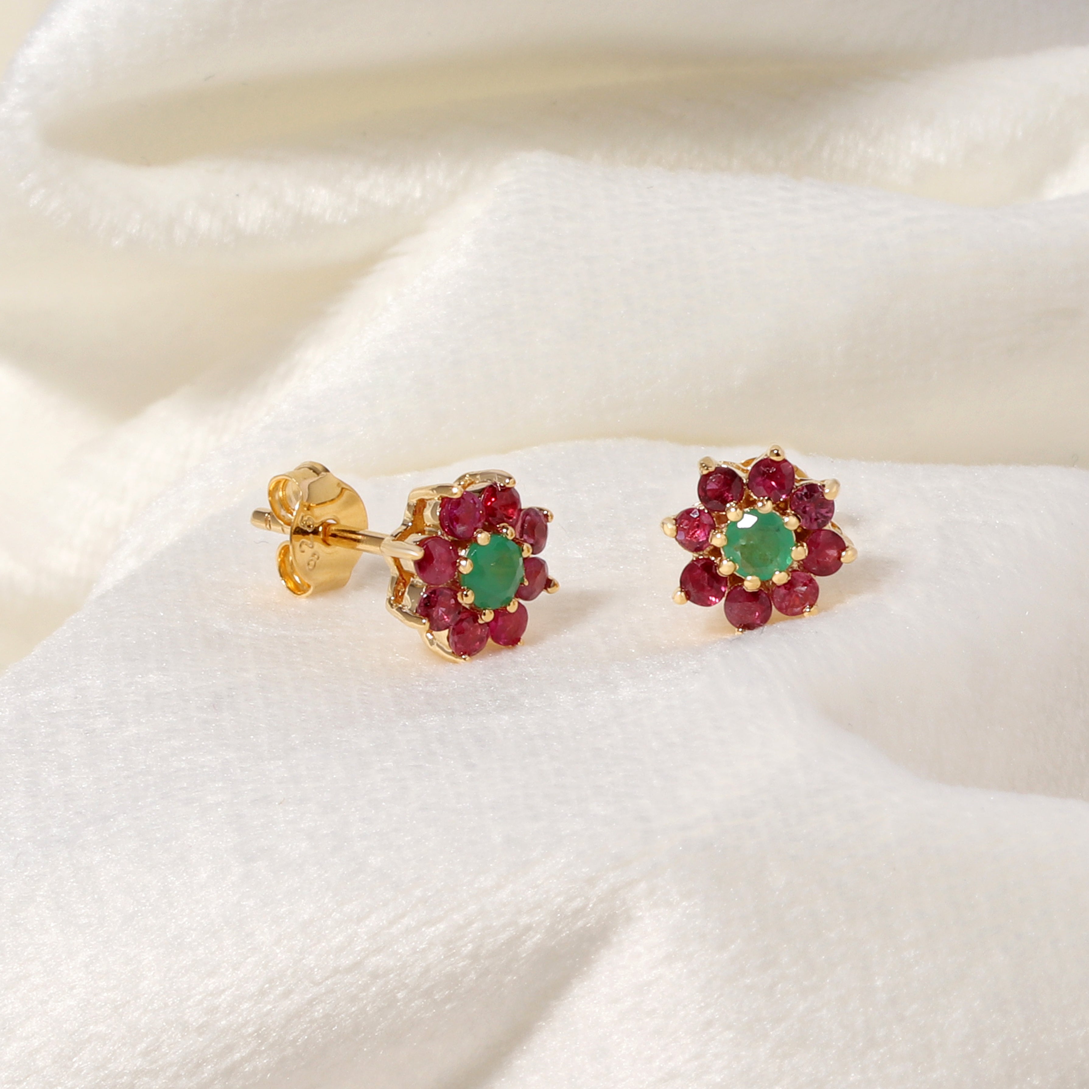 A pair of gold flower-shaped stud earrings with green oval center stones and surrounding rows of red round stones, displayed on a soft white fabric background.